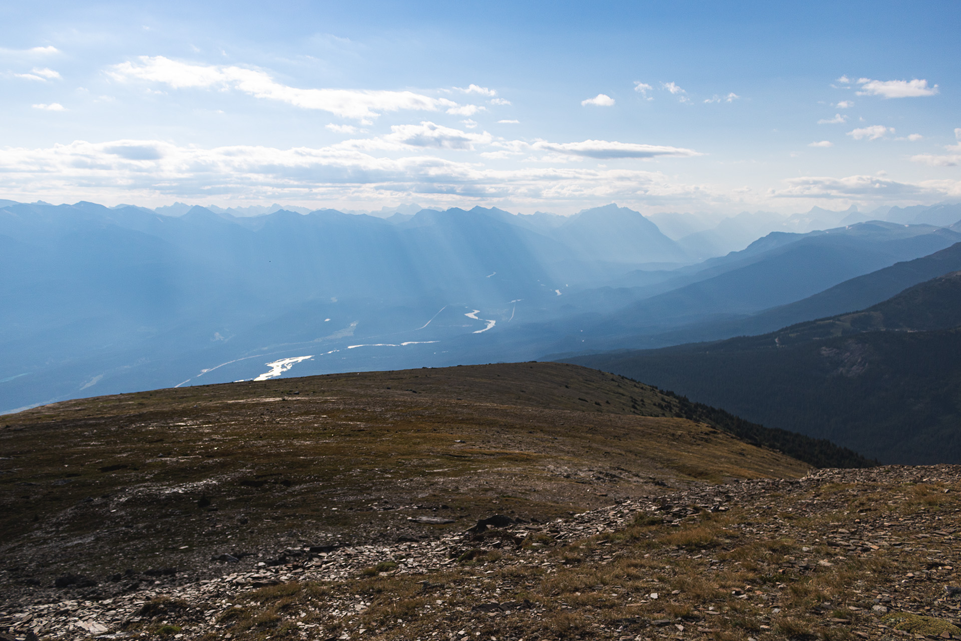 Valley followed by a distant rocky mountain range and a slightly cloudy sky with rays of light passing through it, seen from the top of a mountain.